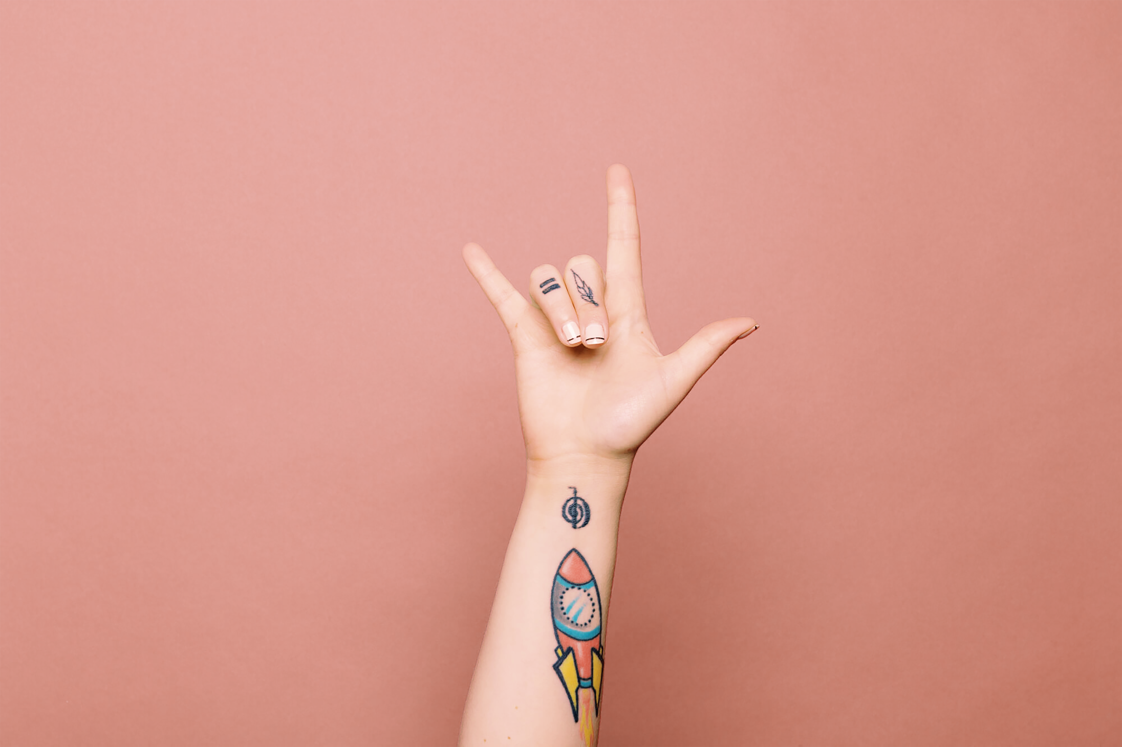 A person's hand and forearm are raised against a solid dusty pink background, showing off small tattoos on the fingers and wrist, including a colorful rocket ship design.
